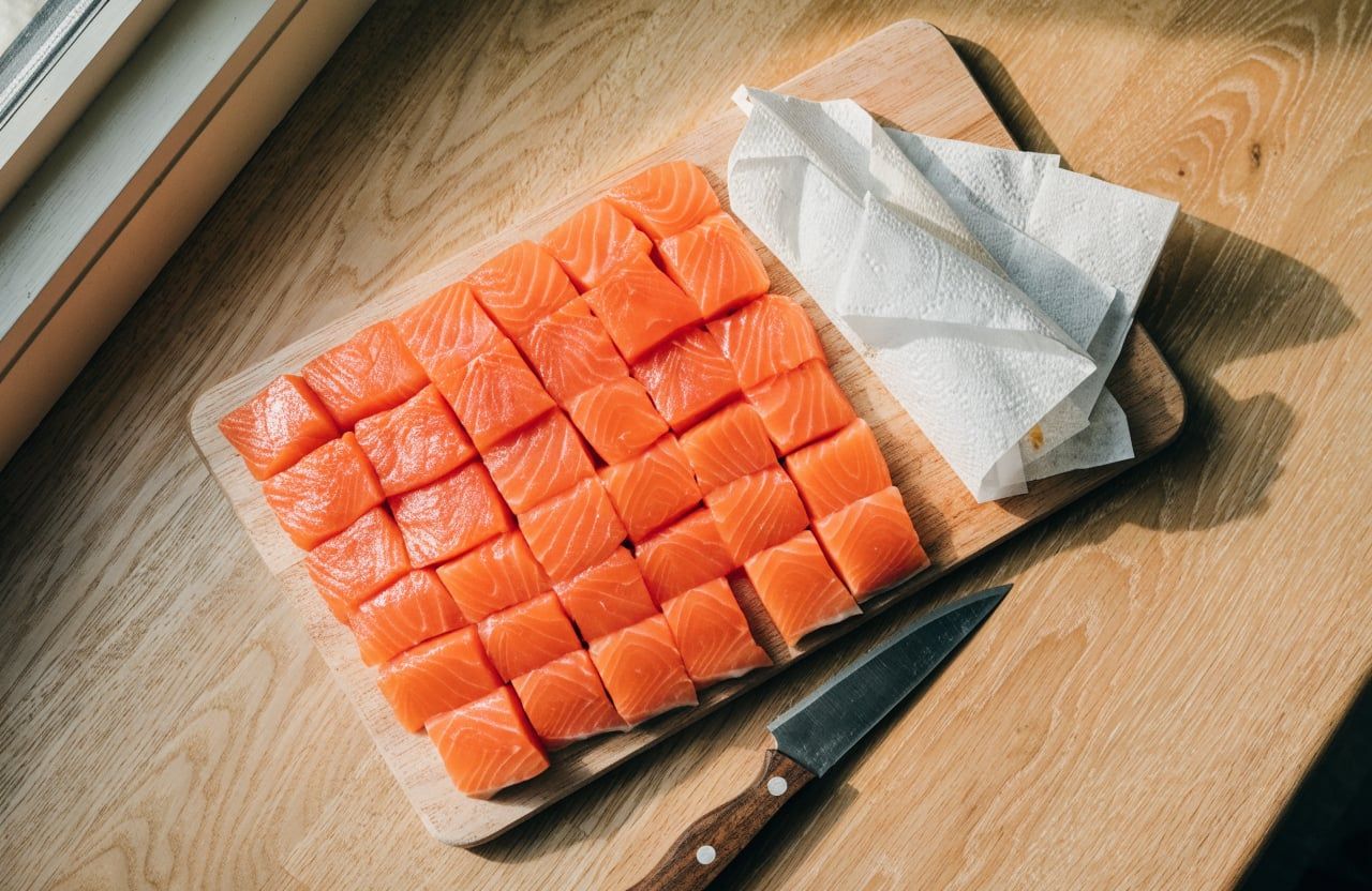 Raw skinless salmon cut into even one-inch cubes on a wooden cutting board with a knife