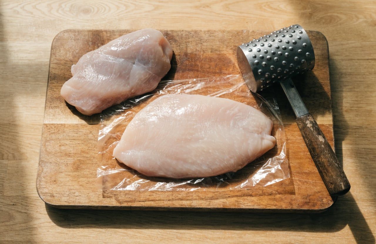 Raw chicken breast pounded to an even thickness between plastic wrap next to a meat mallet
