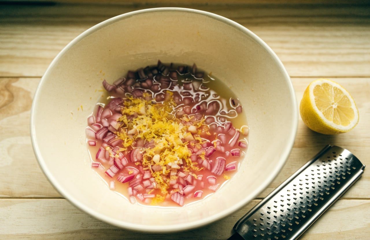 Diced red onion turning pink in lemon juice with zest and grated garlic in a mixing bowl