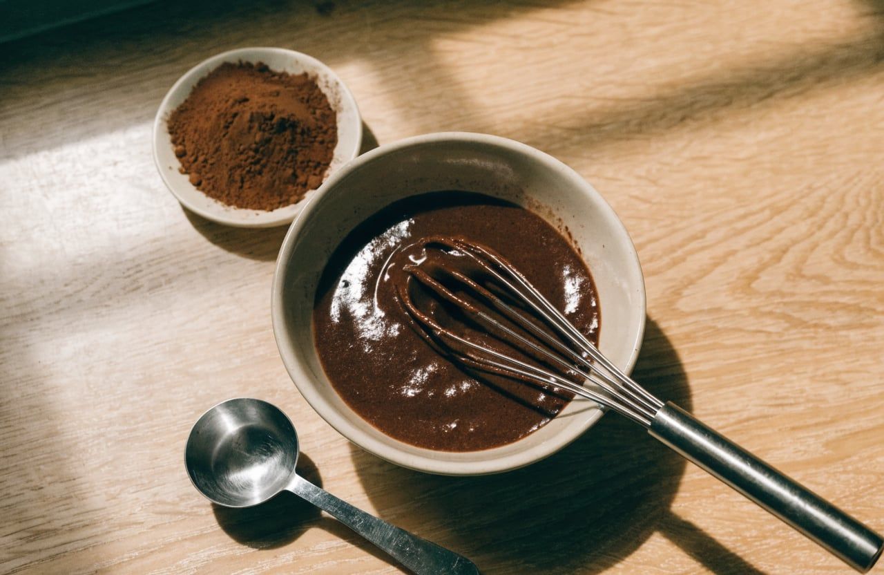 Small bowl with thick, glossy, very dark cocoa and whey paste and a small whisk