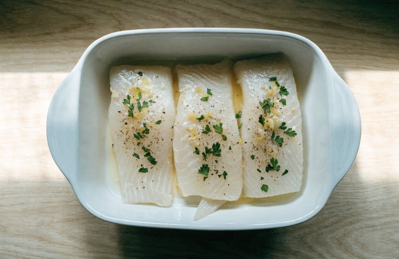 Four raw cod fillets in a baking dish topped with lemon garlic butter before baking