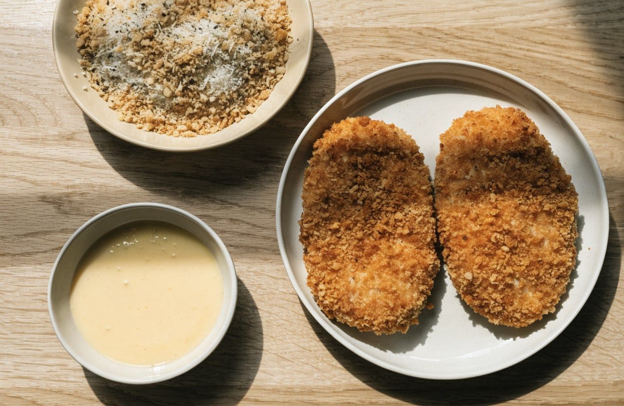 Raw chicken cutlets pressed into golden panko crumbs next to a dish of beaten egg