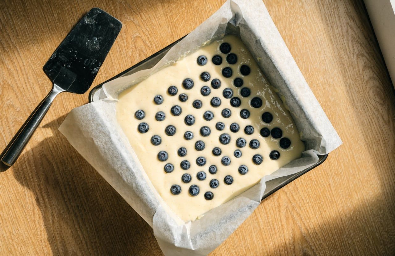 Unbaked ivory batter in a parchment-lined square dish with blueberries pressed into the top