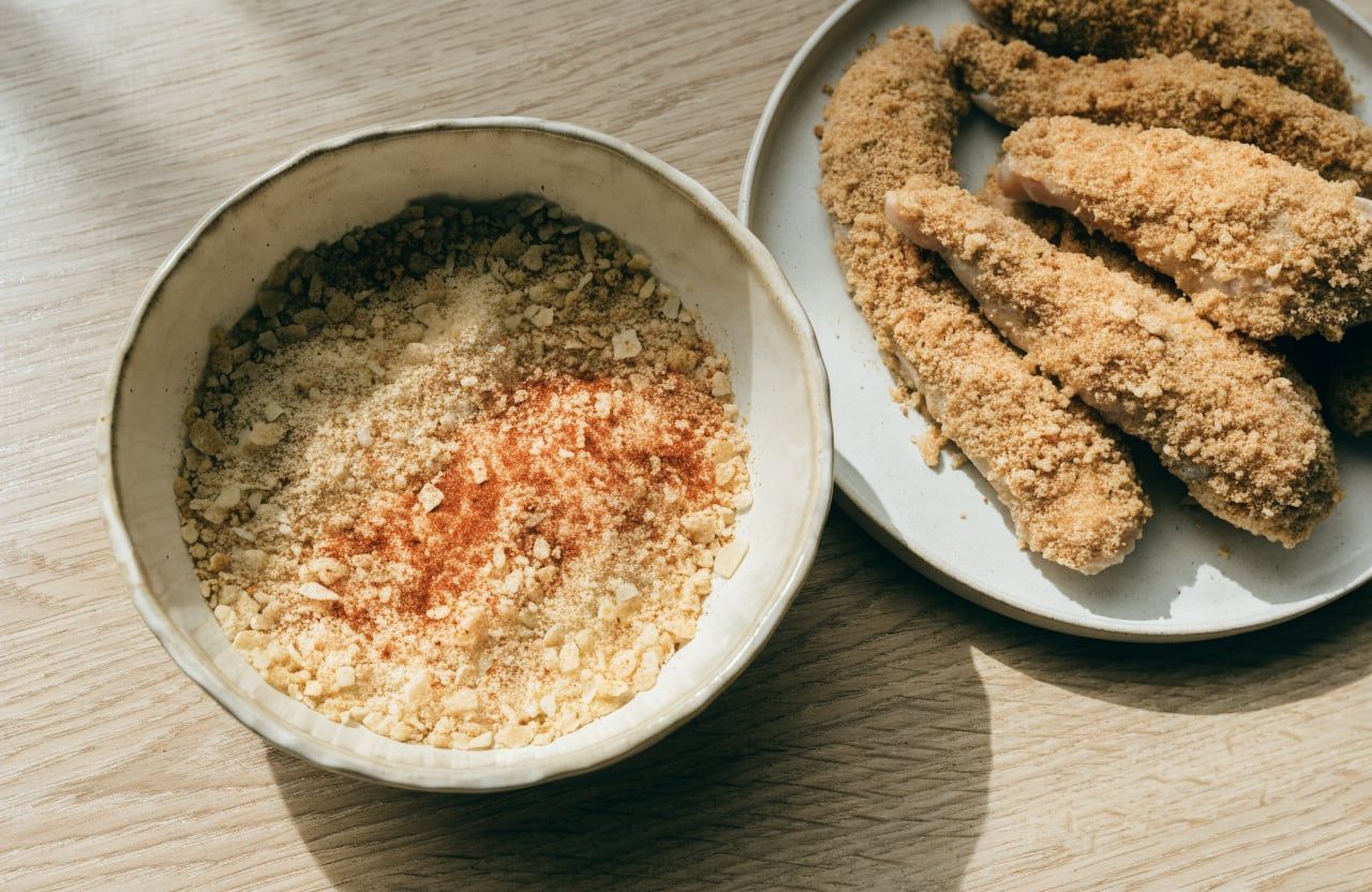 Raw chicken tenders pressed into coarse corn flake crumbs next to a dish of crumbs