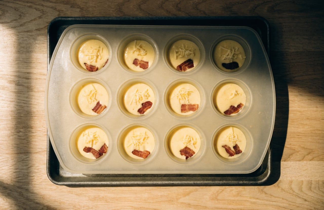 Silicone muffin pan with 12 cups two-thirds full of egg batter topped with shredded Gruyère