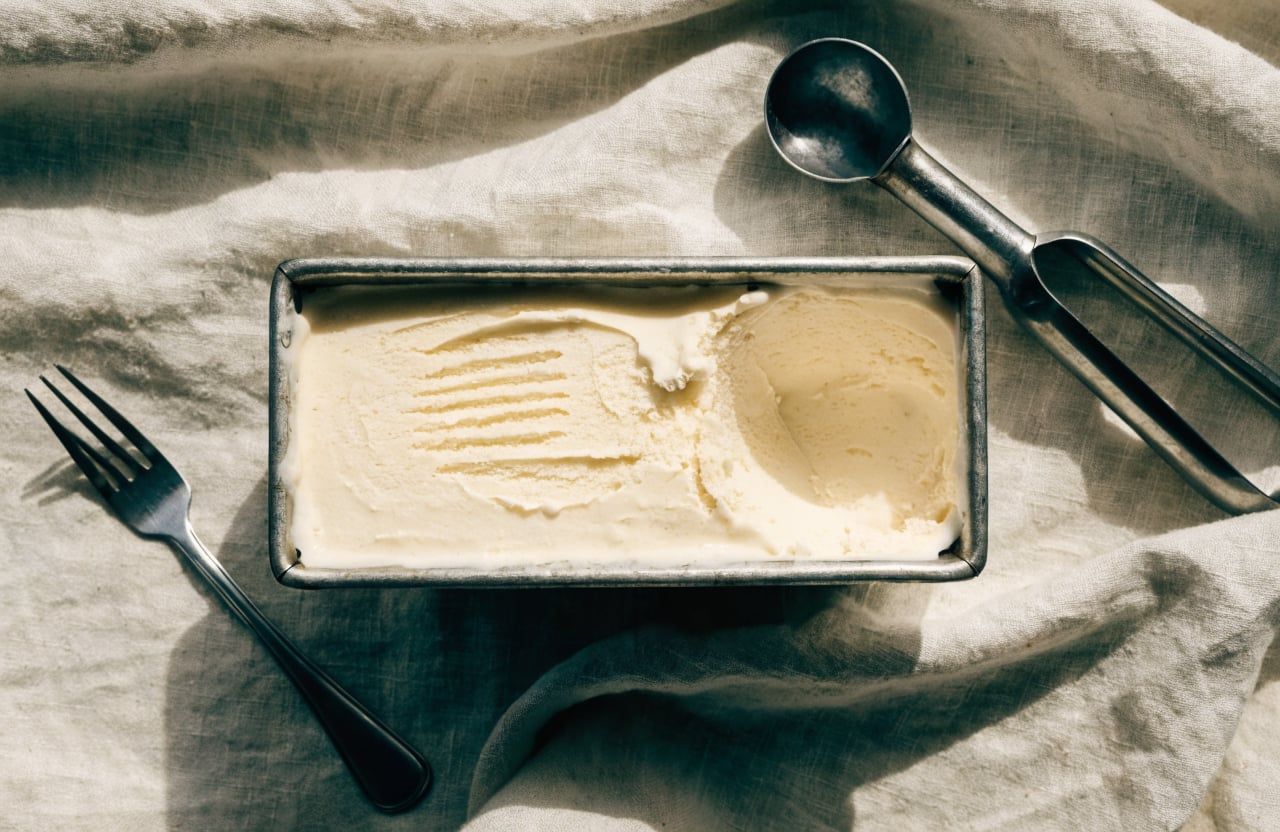 Metal loaf pan of thick, fork-stirred pale ice cream with one scoop taken out