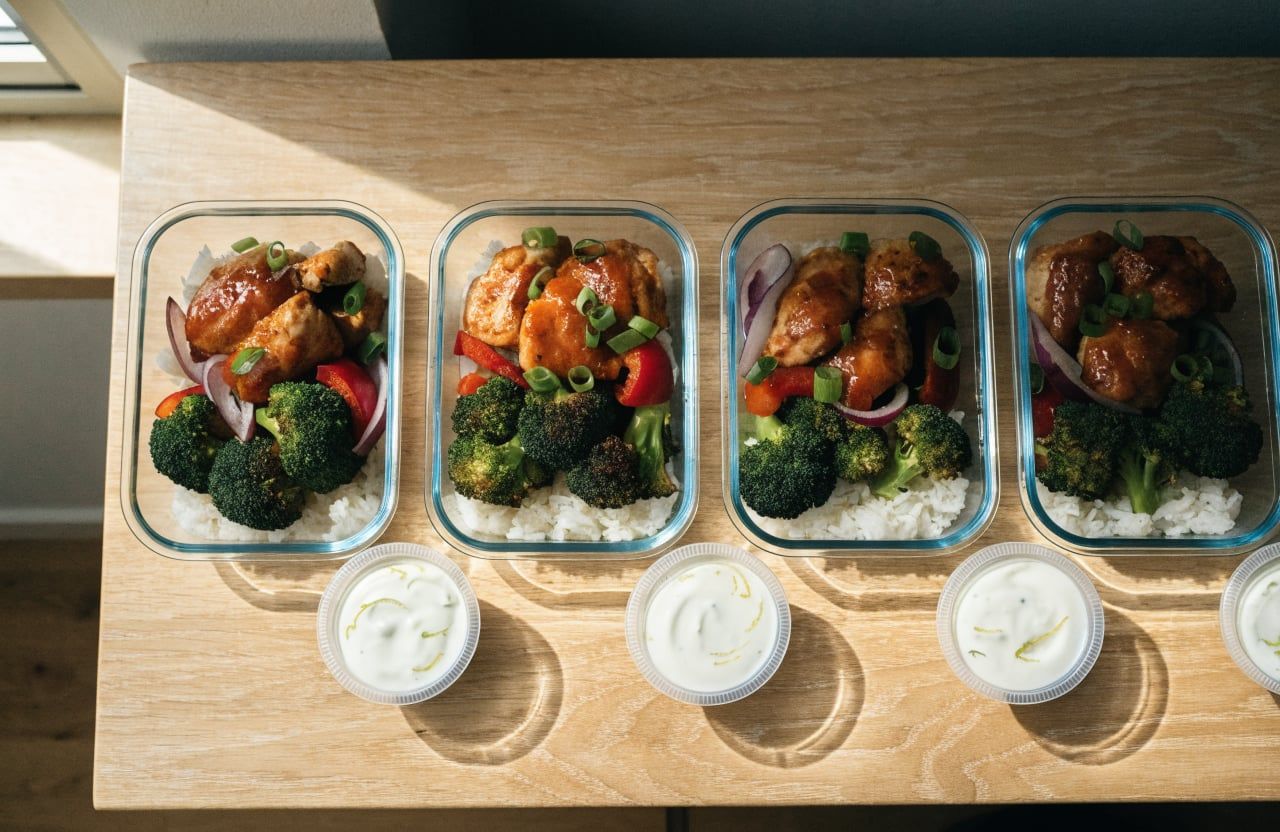 Four meal prep containers with rice, roasted vegetables and glazed chicken, plus yogurt cups