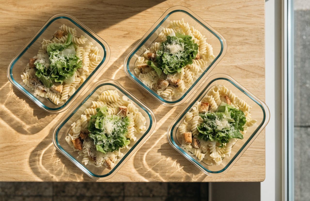 Four meal prep containers of dressed pasta and chicken topped with romaine and parmesan