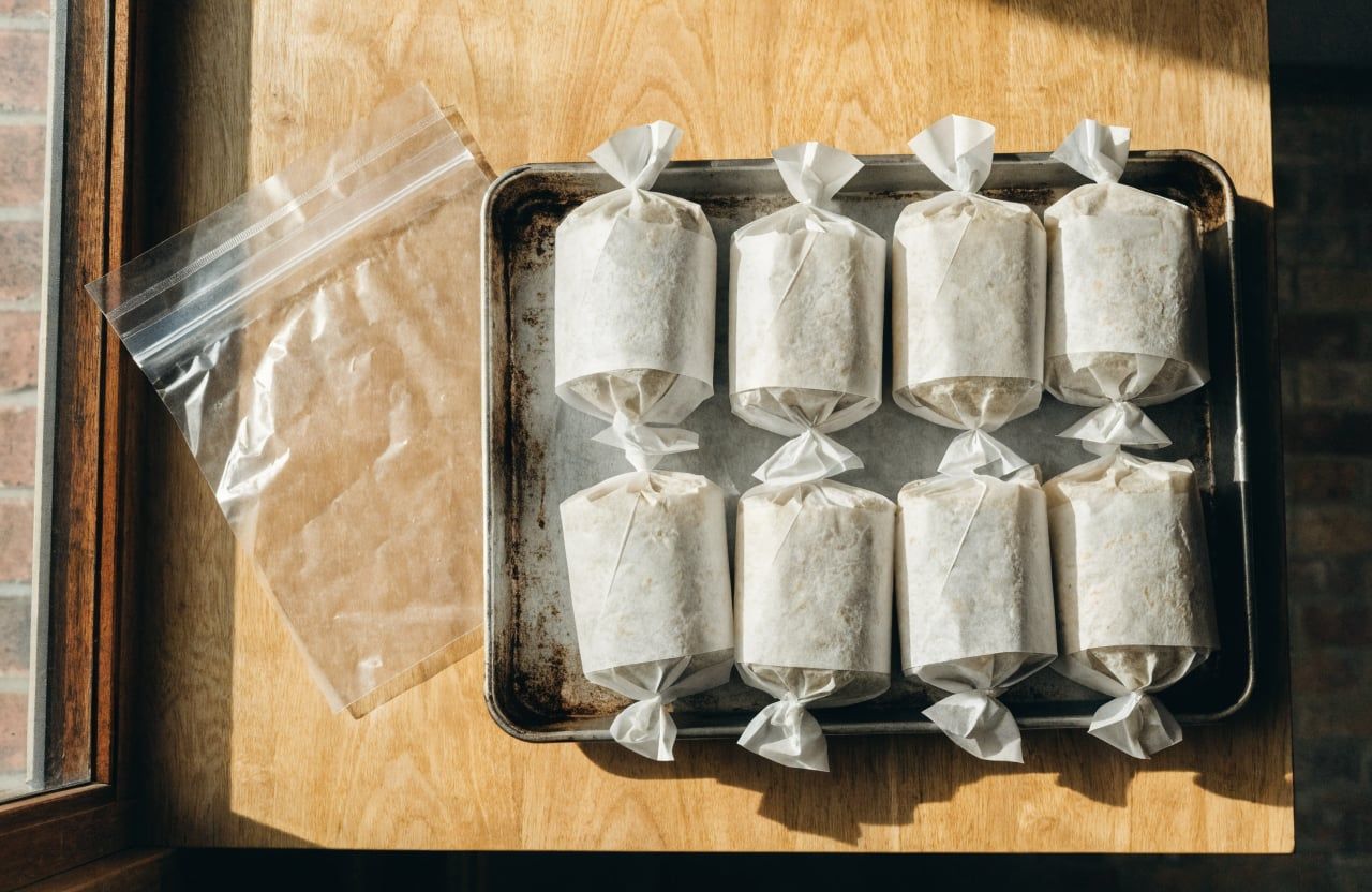 Eight burritos wrapped in parchment with twisted ends lined up on a baking sheet
