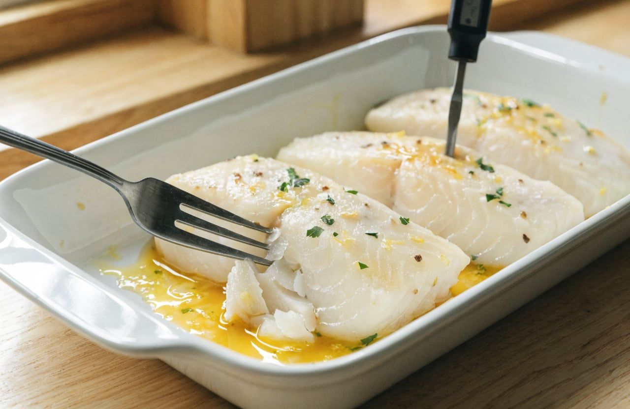 Baked cod fillets flaking under a fork in a baking dish with lemon butter pan juices